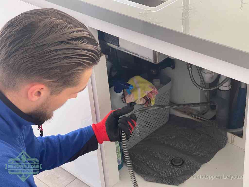 Een monteur voert een flexibele ontstoppingsveer in onder het keukenkastje, langs de sifon, om de gootsteen te ontstoppen in Lelystad