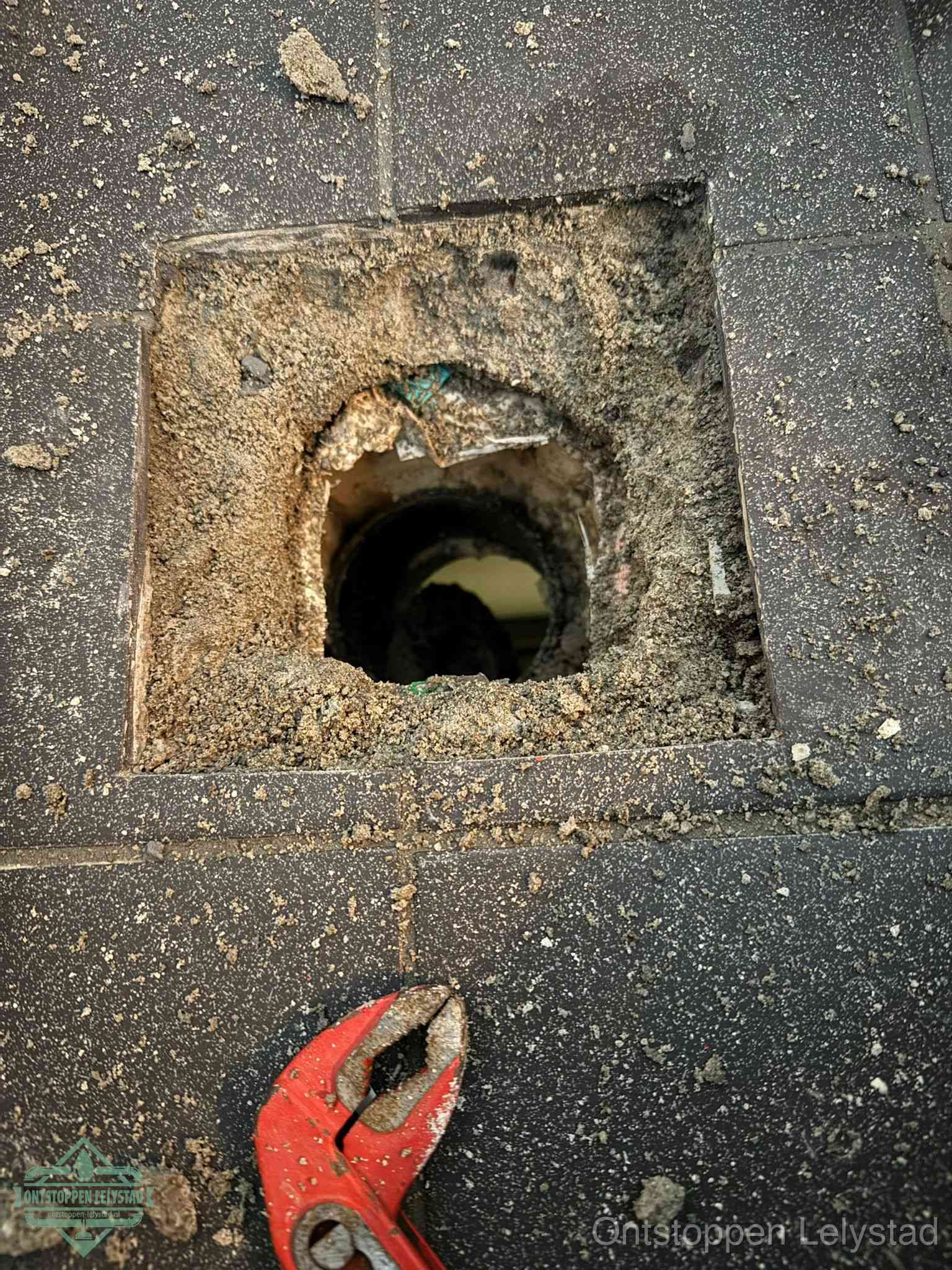 Een geopend rioolputje in een betontegelvloer met losgemaakt rooster en een zichtbare ronde afvoerbuis, het achterhalen waar de afvoerverstopping zit in Lelystad