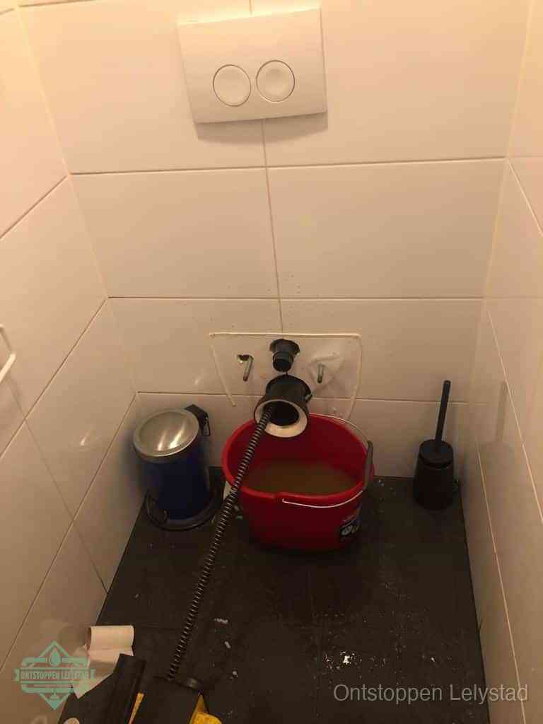 Een ontstopte wand-wc met verwijderde toiletpot waar een ontstoppingsveer in de wanduitgang verdwijnt, het achterhalen waar de verstopping zit in Lelystad