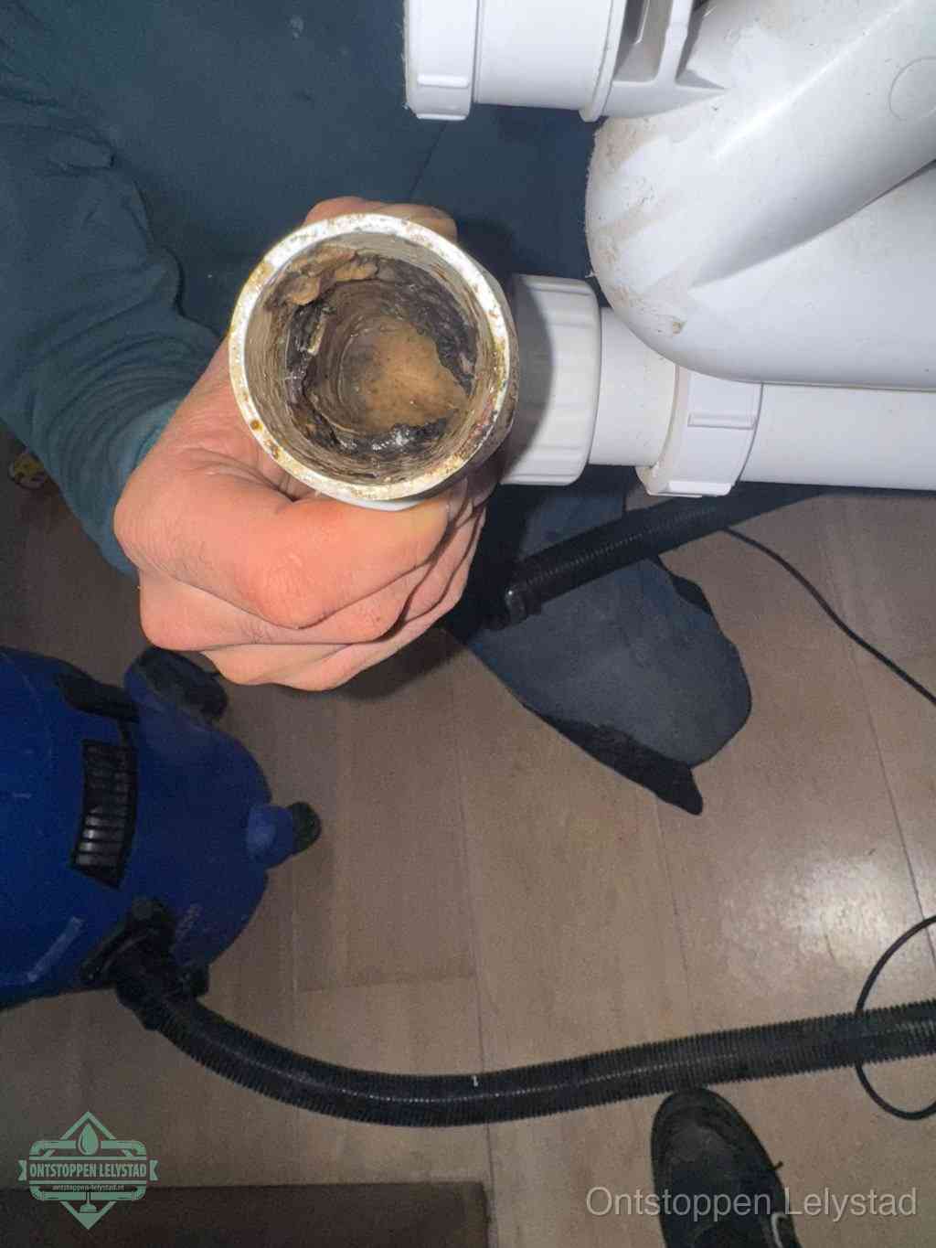 Een hand houdt een losgekoppeld PVC-afvoerbuisstuk vast dat van binnen volledig is dichtgeslibd met vet, vuil en aanslag, de meest voorkomende oorzaak van een verstopte afvoer in Lelystad