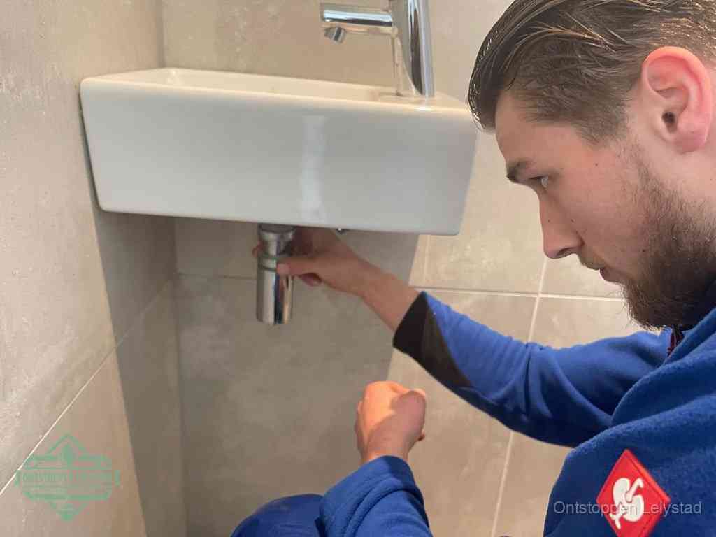 Een monteur draait met de hand de zwanenhalssifon onder een fonteinwasbak in de toiletruimte los om de bron van rioollucht in Lelystad te vinden