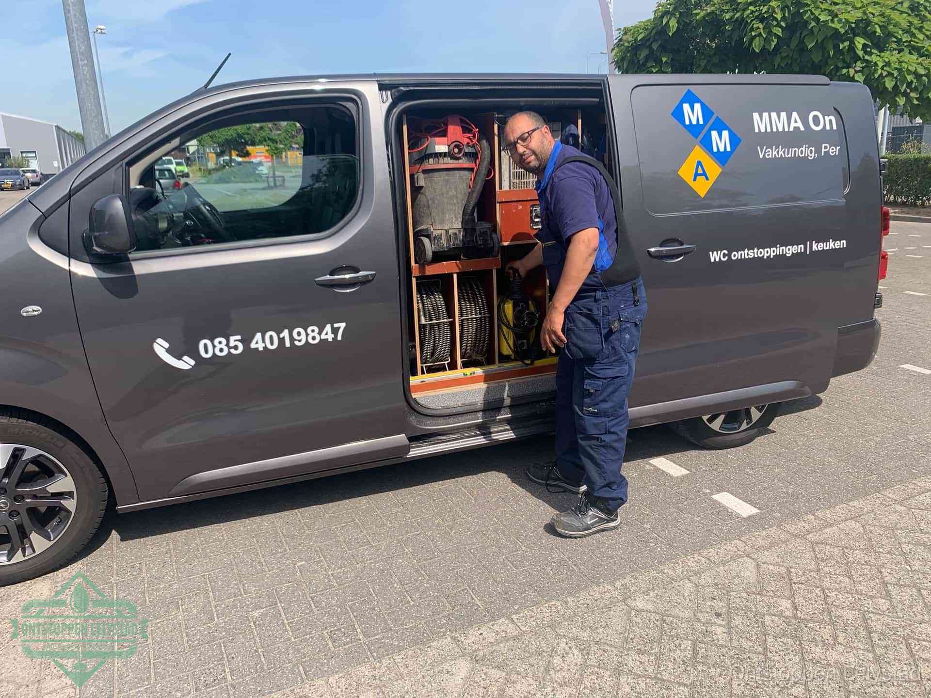 Een monteur bij de open schuifdeur van zijn bedrijfsbus op een parkeerplaats, klaar om de oorzaak van terugkerende afvoerverstoppingen aan te pakken in Lelystad